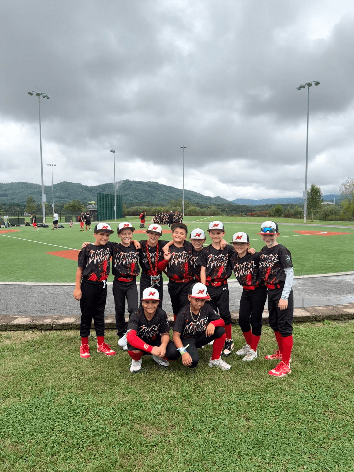 Mamba youth baseball team posing together in black and red custom uniforms beside a baseball field.